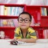 Free A cute child in glasses reading a book, surrounded by colorful shelves. Perfect shot of childhood learning indoors. Stock Photo