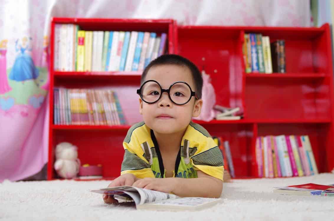 Free A cute child in glasses reading a book, surrounded by colorful shelves. Perfect shot of childhood learning indoors. Stock Photo