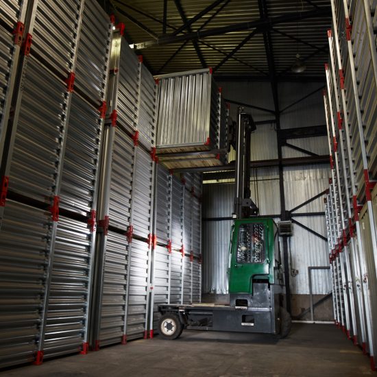 Green powerful forklift truck lowering large metal container in dark cargo storage area