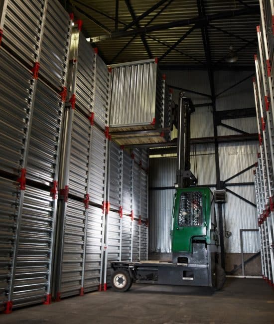 Green powerful forklift truck lowering large metal container in dark cargo storage area